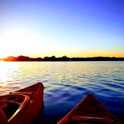 Two orange kayaks on calm water at sunset, with a clear sky and distant trees silhouetted against the horizon.