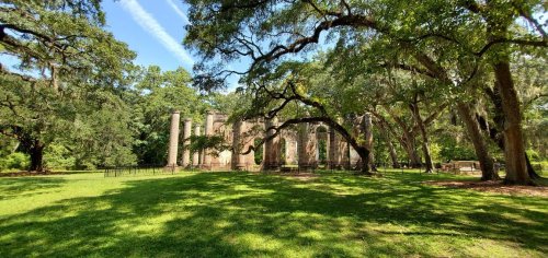 Ruins of a historic structure surrounded by lush greenery and large trees under a clear blue sky.