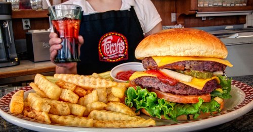 A large cheeseburger with lettuce, tomato, and pickles, served with crinkle-cut fries and a glass of soda.