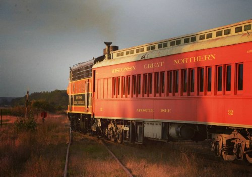 A vintage red train labeled "Wisconsin Great Northern" travels along a scenic, grassy railway at sunset.
