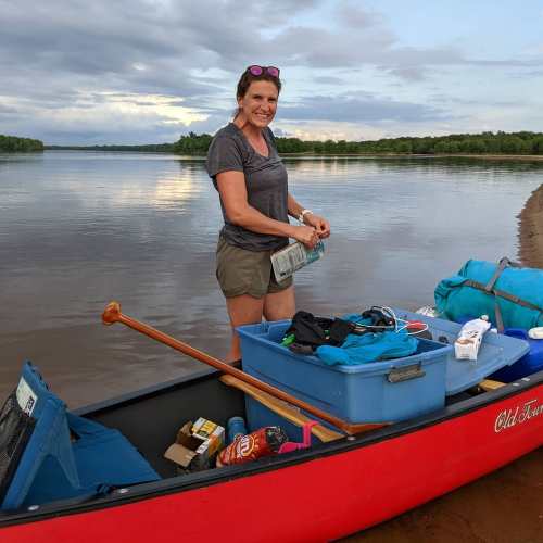 A woman stands by a red canoe on a riverbank, organizing gear and smiling, with a scenic backdrop of water and trees.