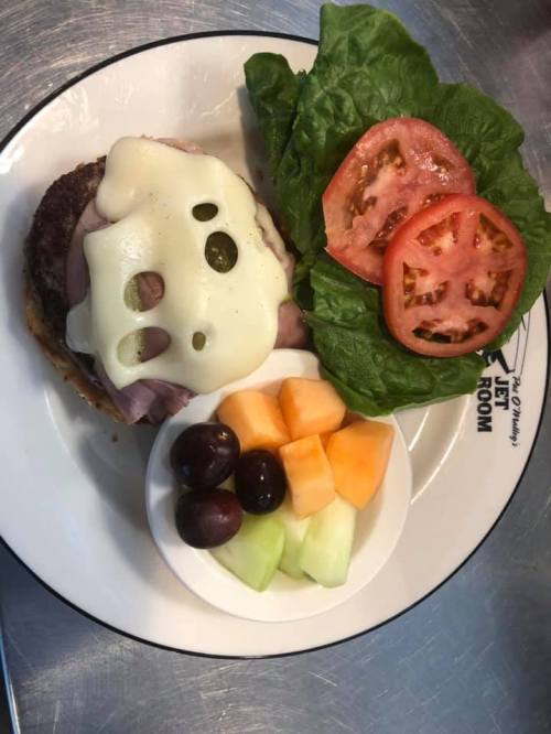 A plate featuring a sandwich with melted cheese, lettuce, tomato, and a side of mixed fruit.