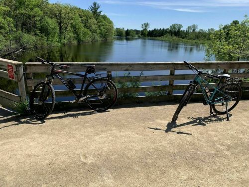 Two bicycles parked on a sandy path beside a calm lake, surrounded by lush greenery and a clear blue sky.