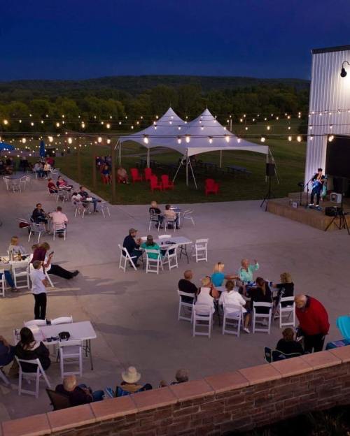 Outdoor event at dusk with string lights, tents, and people enjoying music and dining in a scenic setting.