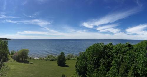 A panoramic view of a calm lake under a clear blue sky, surrounded by lush green trees and grass.