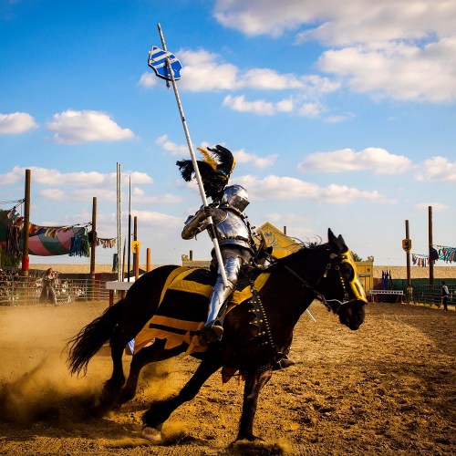 A knight in shining armor rides a horse, wielding a lance, in a dusty arena under a blue sky with clouds.