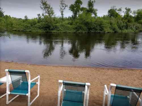 Chairs on a sandy beach facing a calm river surrounded by lush greenery.