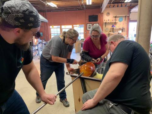 Four people collaborate on glassblowing, shaping a glowing orange piece in a workshop setting.
