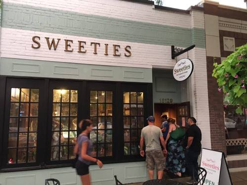 A group of people stands outside a shop called "Sweeties," with a sign indicating it's open.