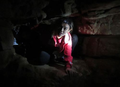 A person in a red jacket crawls through a dark cave, illuminated by a headlamp, with rocky walls surrounding them.