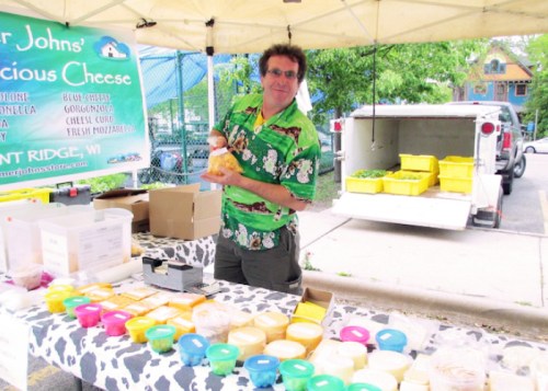 A man in a Hawaiian shirt stands at a market stall displaying various cheese products and colorful containers.