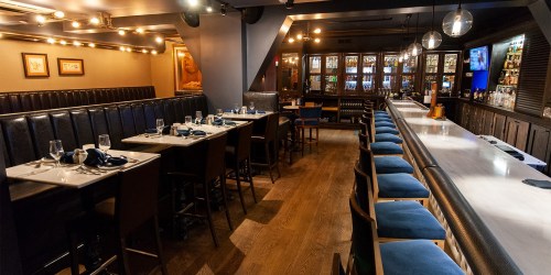 A stylish restaurant interior featuring a long bar, dining tables, and elegant lighting.