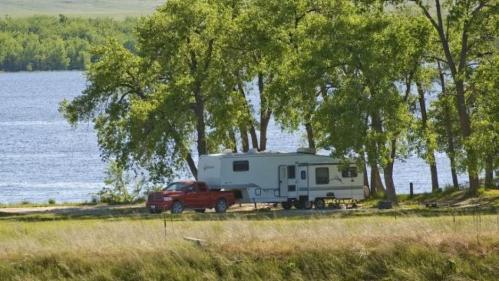 A red truck towing a camper parked by a serene lake, surrounded by green trees and grassy land.