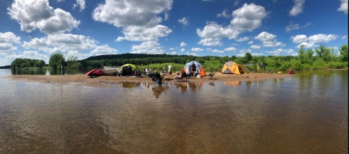 A sandy riverbank with colorful tents, people gathered, and lush greenery under a bright blue sky with fluffy clouds.