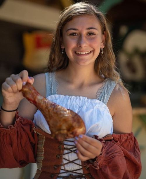 A smiling young woman in a historical costume holds a large cooked turkey leg.