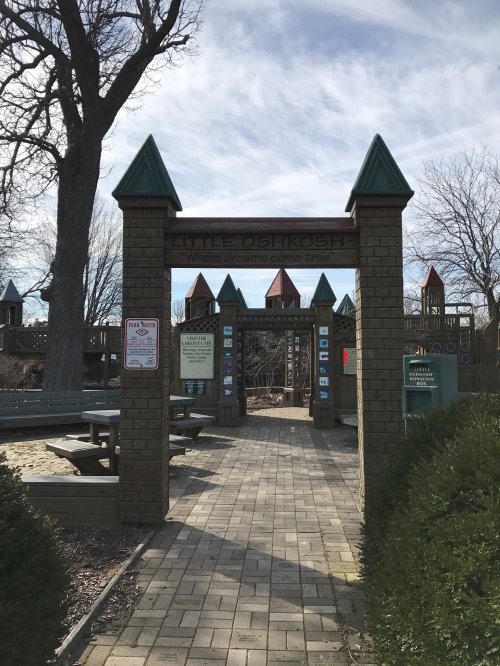 Entrance to a playground with a sign reading "Little Obikosh," surrounded by trees and colorful play structures in the background.