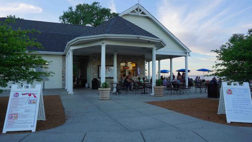 A lakeside café with outdoor seating, umbrellas, and menu boards outside, set against a scenic sky.