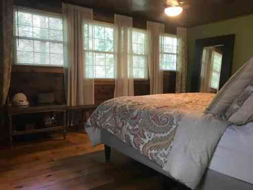 Cozy bedroom with a patterned bedspread, large windows, and wooden accents, featuring a mirror and soft curtains.
