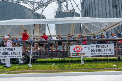 A crowd of people seated under a tent, enjoying food and drinks near silos at a community event in Seymour, WI.