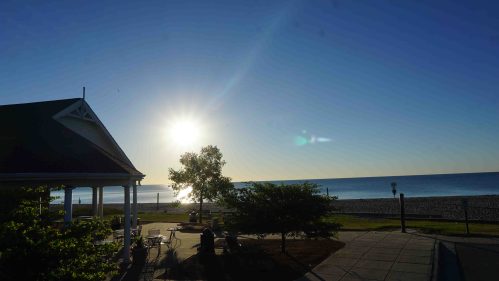 Sunrise over a calm beach, with a gazebo and trees in the foreground, casting long shadows on the pavement.