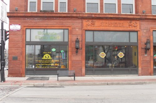 Exterior of a brick building featuring "Winnetka Cheer Mart" and "Sternmeyer" signs, with large windows and a bench outside.
