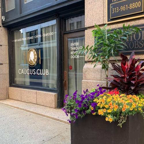 Exterior of Caucus Club restaurant with a sign, large window, and colorful flower planter.