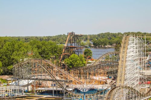A view of a roller coaster under construction near a lake, surrounded by trees and a clear blue sky.