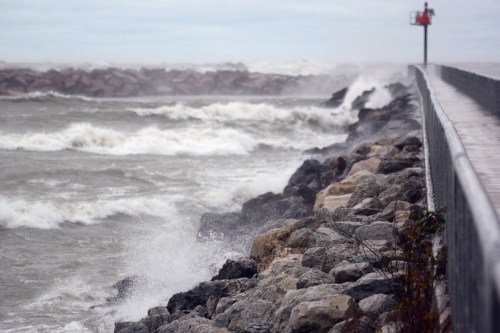 Waves crash against a rocky shoreline with a pier and a red lighthouse in the background on a stormy day.