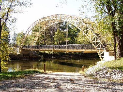 A yellow arch bridge spans a calm river, surrounded by trees and greenery on a sunny day.