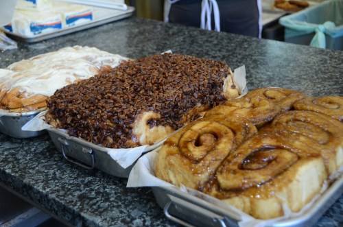 Three trays of freshly baked cinnamon rolls: one with cream cheese frosting, one with chocolate topping, and one glazed.