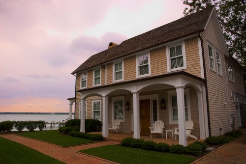 A charming two-story house with a porch, set by a waterfront, surrounded by green grass and a cloudy sky.