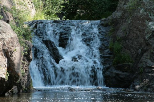 A serene waterfall cascading over rocky terrain, surrounded by lush greenery and calm water below.