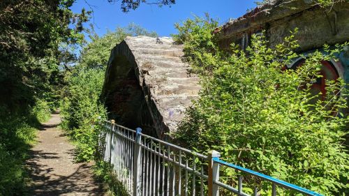 Overgrown path beside a weathered, curved concrete structure, surrounded by lush greenery and blue sky.