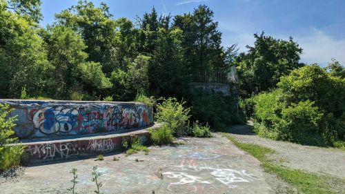 Overgrown area with a circular, graffiti-covered structure and a path leading through dense greenery.