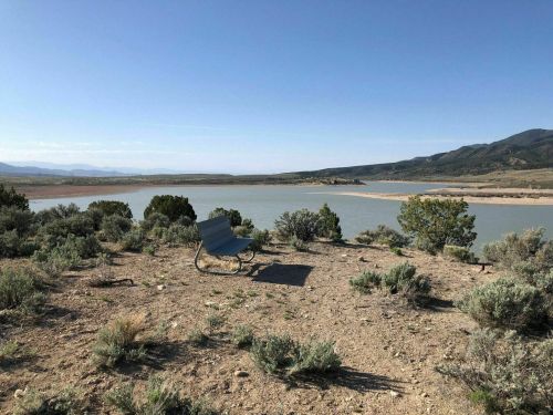 A lone chair sits on a rocky shore overlooking a calm lake surrounded by shrubs and distant mountains.