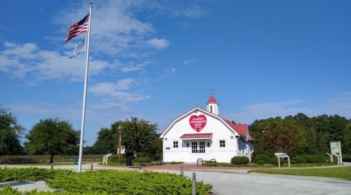 A white building with a red roof and sign, surrounded by greenery, under a blue sky with clouds and an American flag.