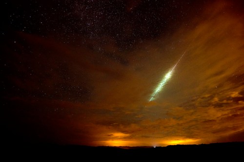 A night sky filled with stars and clouds, featuring a bright meteor streaking across the horizon.