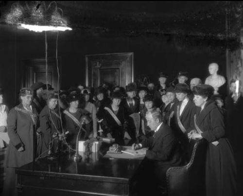 A historic gathering of women in hats observing a man signing documents at a desk in a formal setting.