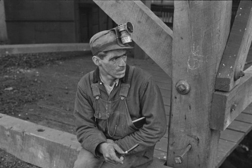 A miner sitting on a wooden structure, wearing a cap with a lamp, looking thoughtfully to the side.