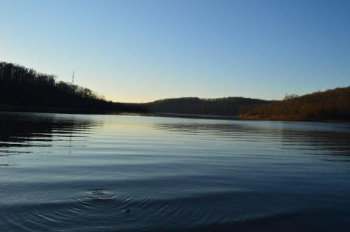 A serene lake at sunset, with calm waters reflecting the sky and surrounding hills.