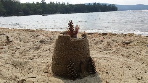 A sandcastle on a beach, decorated with pinecones, with a lake and trees in the background.