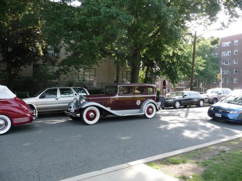 A vintage maroon and white car parked on a tree-lined street with several other cars in the background.