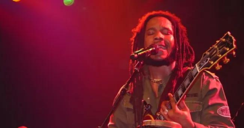 A musician with dreadlocks sings passionately into a microphone while playing guitar on stage, illuminated by colorful lights.
