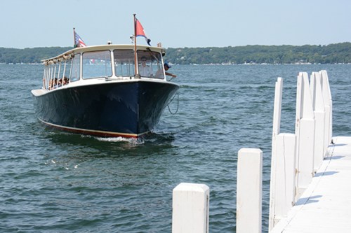 A boat approaches a wooden dock on a sunny day, with calm water and green trees in the background.