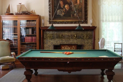 A vintage billiard table in a room with a fireplace, bookshelves, and a large painting on the wall.