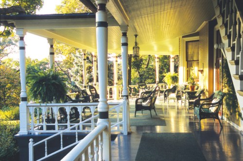 A spacious porch with wicker chairs and plants, surrounded by greenery and bathed in warm sunlight.