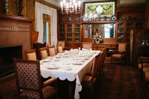 A vintage dining room with a long table set for a meal, surrounded by ornate wooden furniture and decorative glass cabinets.