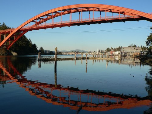 A vibrant orange bridge arches over calm water, reflecting its colors, with a small harbor and trees in the background.