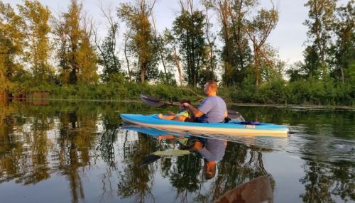 A person kayaking on calm water, surrounded by trees, with a serene sunset reflecting on the surface.