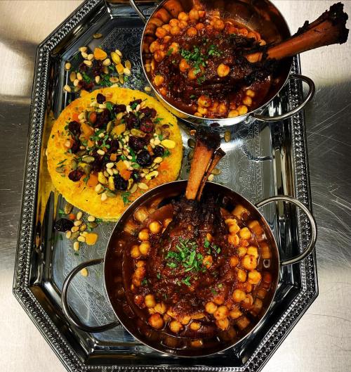 Two bowls of chickpea stew with lamb shanks, garnished with herbs, served on a decorative tray with a colorful flatbread.